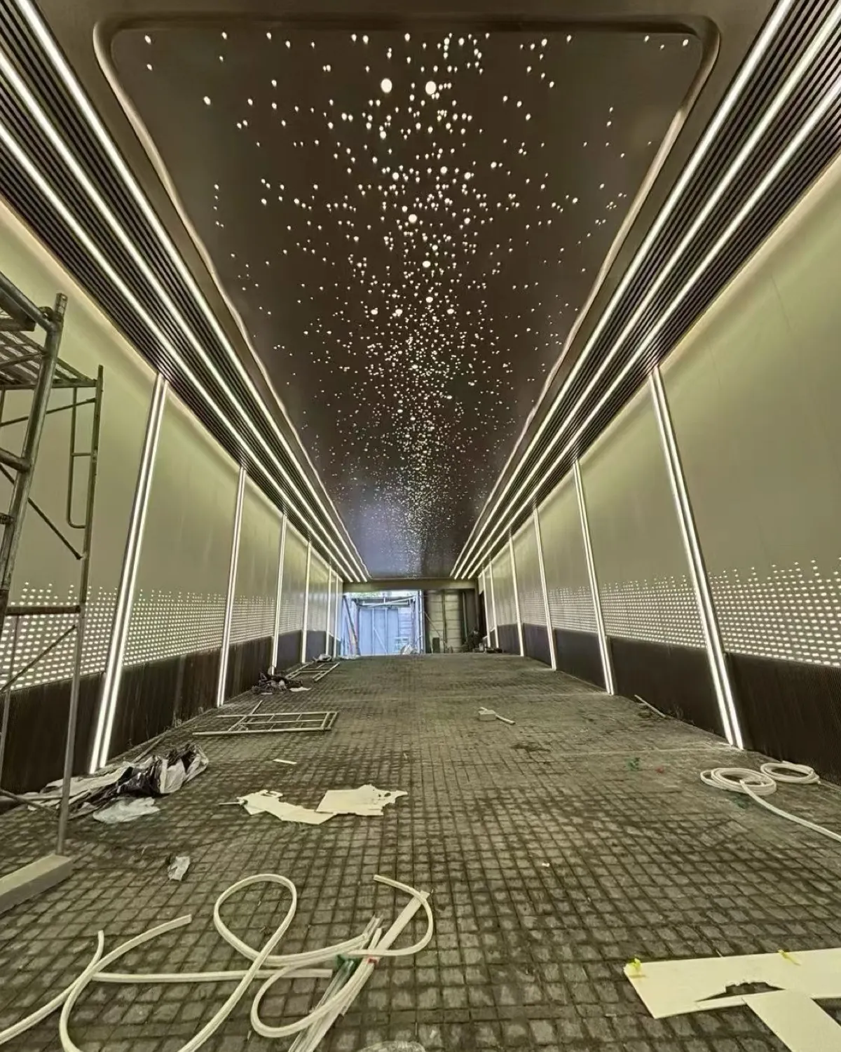 Installation-stage corridor with linear lights, perforated ceiling and wall areas, scaffolding and unfinished flooring