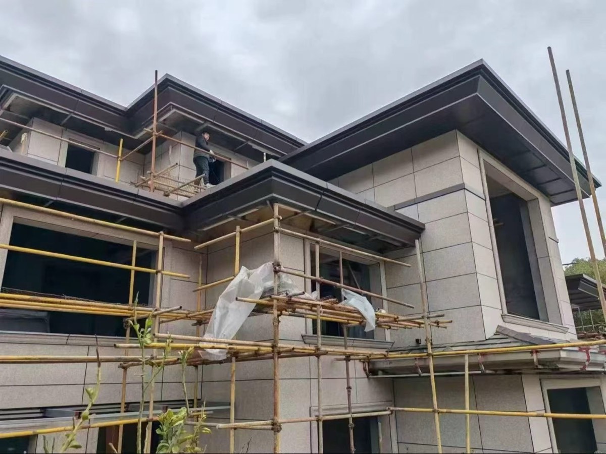 Dark roof-edge fascia and soffit cladding above pale walls, with scaffolding and protective sheeting below.