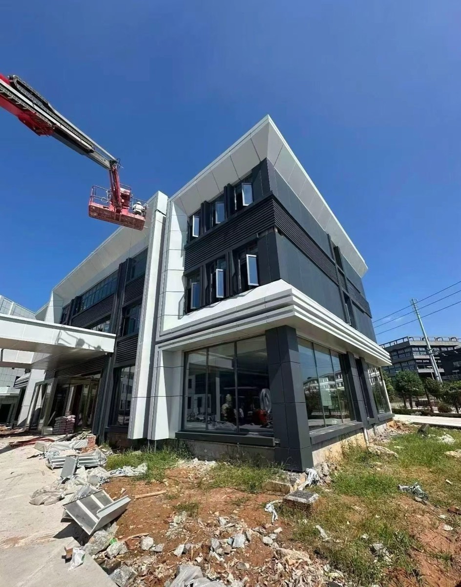 White roof-edge cladding frames a dark angular facade, with windows and a red access platform above unfinished ground.